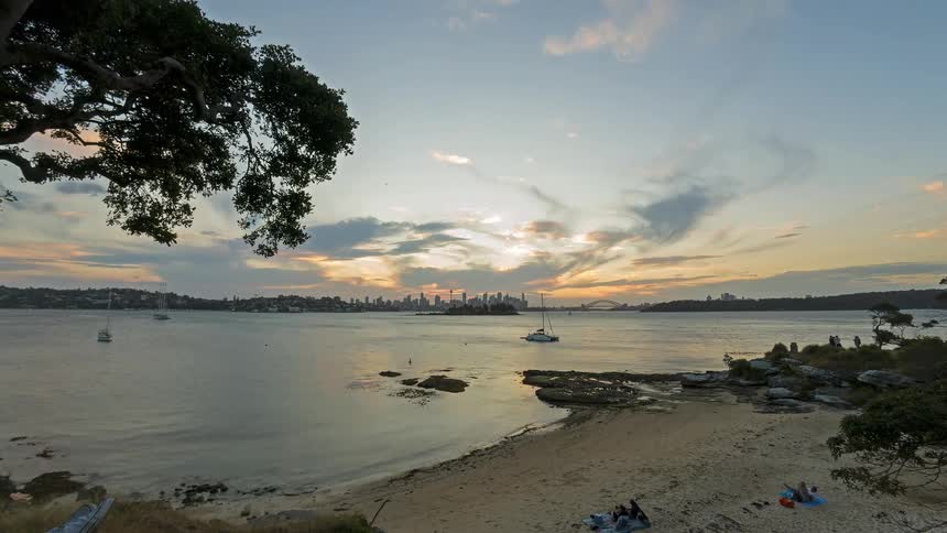 This timelapse captures a golden sunset over a harbor with the sun dipping behind a distant city skyline and its reflection shimmering on the water as dusk settles wispy clouds stretch across the sky while the city lights begin to twinkle and the beac