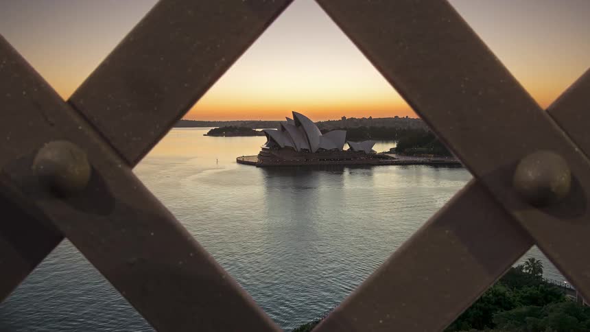 A time lapse view looking through the metal lattice of a bridge shows the sun rising behind a large white opera house on a harbor with boats moving across the calm water as the sky changes from purple to orange