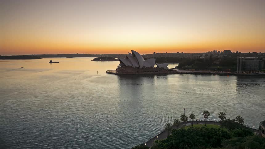 A time lapse shows the sun rising over the sydney opera house and a harbor as boats move across the water and the sky changes from dark purple to bright orange