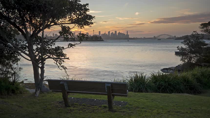 A wooden bench sits on a grassy hill overlooking a calm harbor at sunset with a city skyline and a large bridge visible in the distance under an orange sky