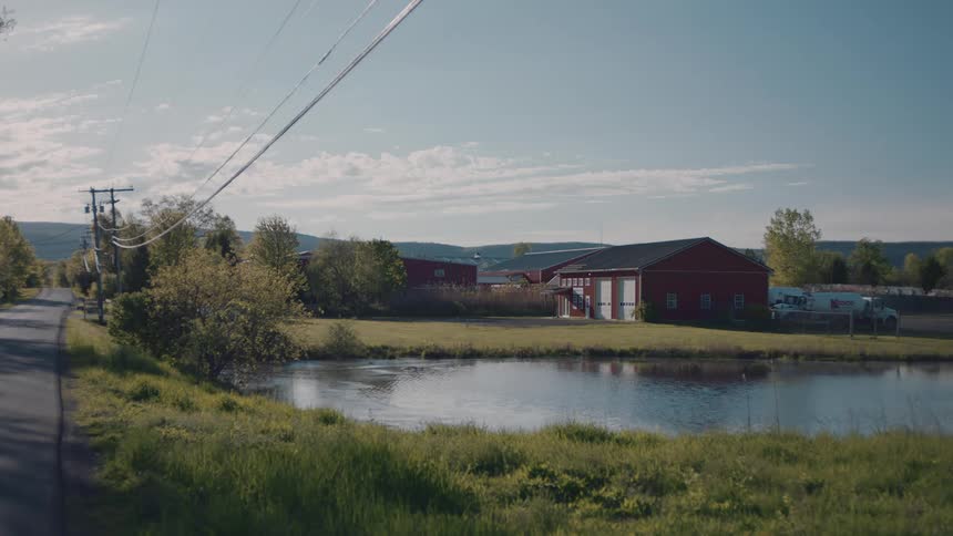 A red barn with a dark roof stands near a calm pond and a paved road with power lines under a blue sky with light clouds during a clear day in a rural setting