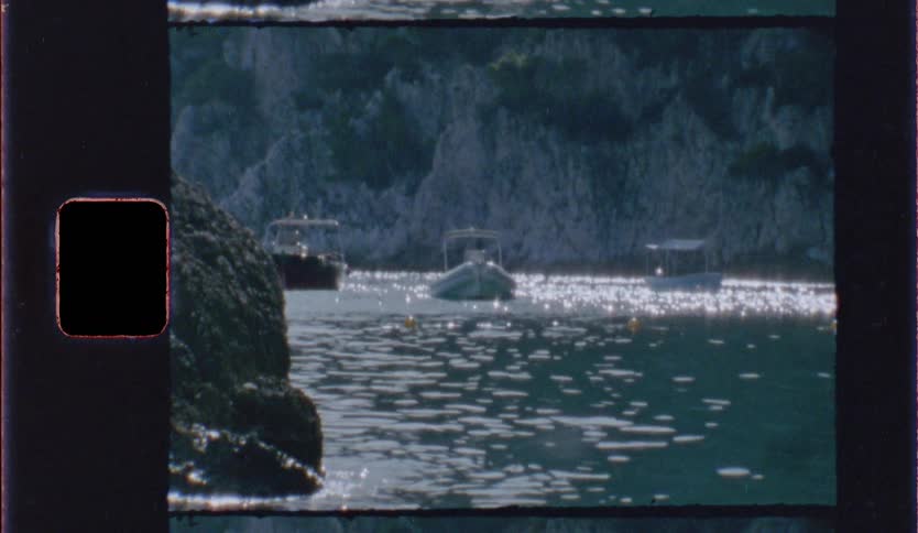 Vintage film footage shows three small boats anchored on calm water near a rocky cliff with bright sunlight reflecting off the ripples creating a shimmering effect