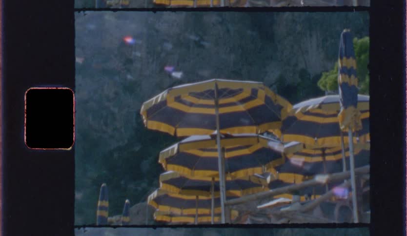 A collection of yellow and black striped beach umbrellas clustered together against a rocky coastal backdrop it is filmed with a vintage aesthetic complete with film grain light leaks and a visible film sprocket hole on the left