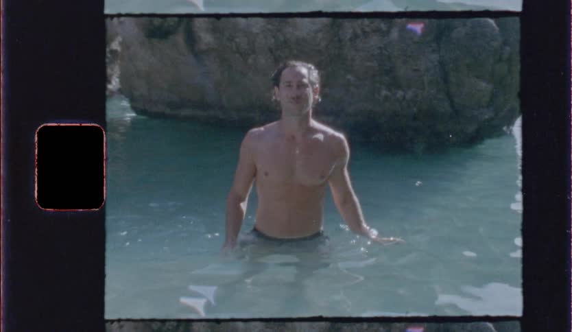 A shirtless young man walking in a pond in the wilderness at midday with a large rock behind him