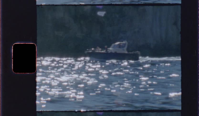 A small motorboat moves across shimmering blue water in front of a dark rocky cliff in a vintage film style