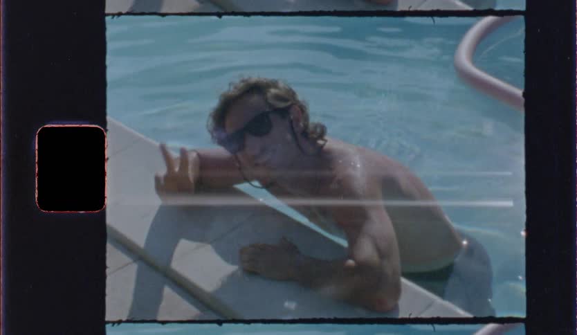 A man wearing sunglasses relaxes in a swimming pool leaning on the edge and flashing a peace sign framed in a vintage filmstyle border