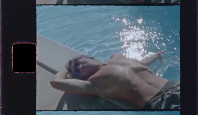 A man lies shirtless waving on a lounge chair beside a sparkling blue pool sunbathing under bright daylight in a vintagestyle frame