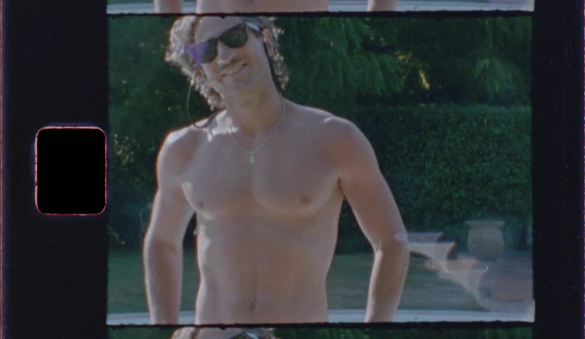 Shirtless man wears sunglasses outdoors with a garden in the background