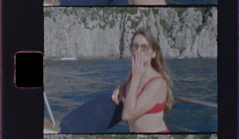 A woman in a red bikini and sunglasses smiles and blows a kiss while sitting on a boat holding a dark blue cushion under her arm with rocky cliffs and blue water in the background framed in a vintage filmstyle border