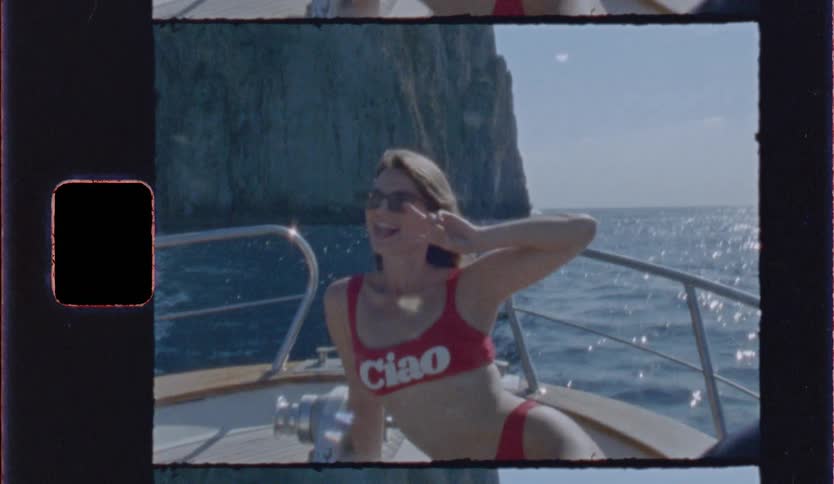 A woman in a red bikini relaxing on a boat as it cruises past towering coastal cliffs the footage has a vintage grainy film aesthetic complete with visible sprocket holes and a nostalgic summer atmosphere