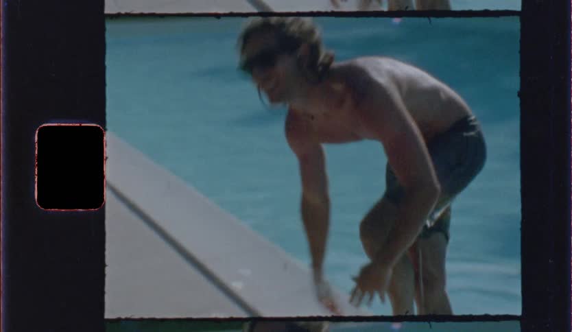 A man wearing sunglasses and swim trunks climbs out of a blue swimming pool and walks along the edge on a sunny day in a vintage film style