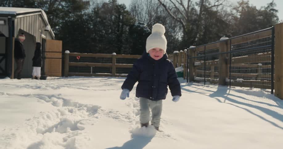 Little boy walking through the snow in his yard as his parents stand near the house