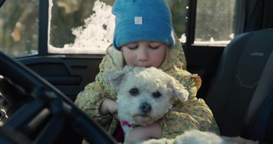 A young girl wearing a blue knit hat and a yellow floral jacket smiles and looks at the camera while hugging a small white dog in the front seat of a vehicle on a bright day
