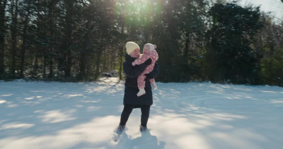 An adult in a long dark coat lifts a bundled child in pink winter clothing into the air and spins the child around in play in a snowy clearing with sunlight streaming through tall trees in the background