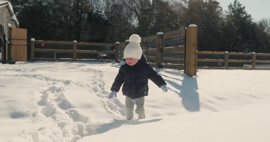 A young child wearing a blue coat and a white hat walks through deep snow and smiles in a sunny yard with a wooden fence