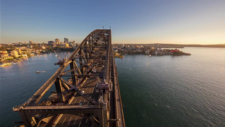 A high angle view shows vehicles driving across a large steel bridge over a harbor during a golden sunset with a city skyline and boats on the water in the background