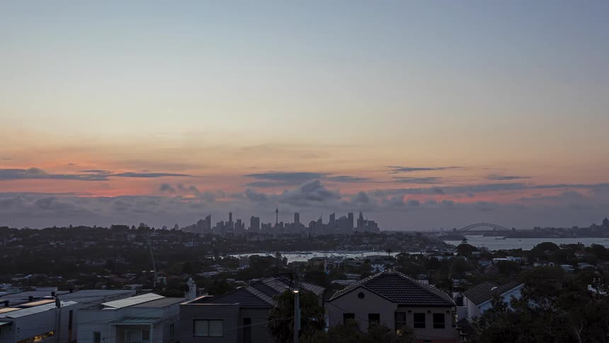 Traffic moves in a busy neighborhood on the coast as boats sail on the water and under a bridge with the downtown skyline on the horizon as the sun sets