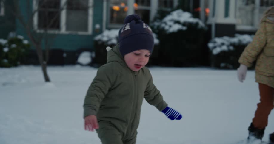 A toddler walking on the snow beside an excited little girl on the front lawn at midday
