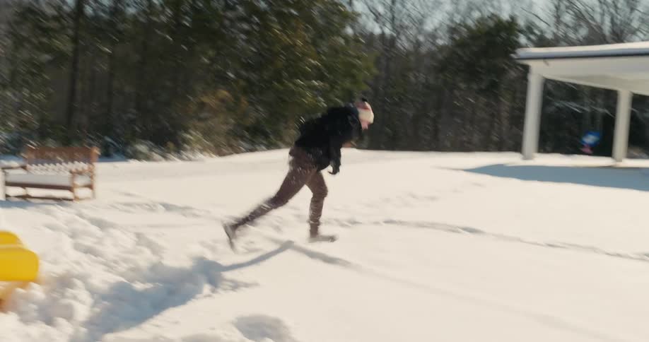 A man wearing a black jacket and beanie slides down a yellow slide into the snow and runs toward the camera laughing in a sunny backyard with a wooden playground and a white house in the background