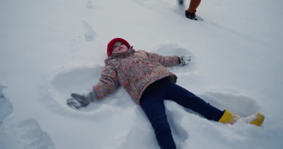 A young child wearing a red hat and a floral jacket lies in thick white snow while moving their arms and legs to create a snow angel