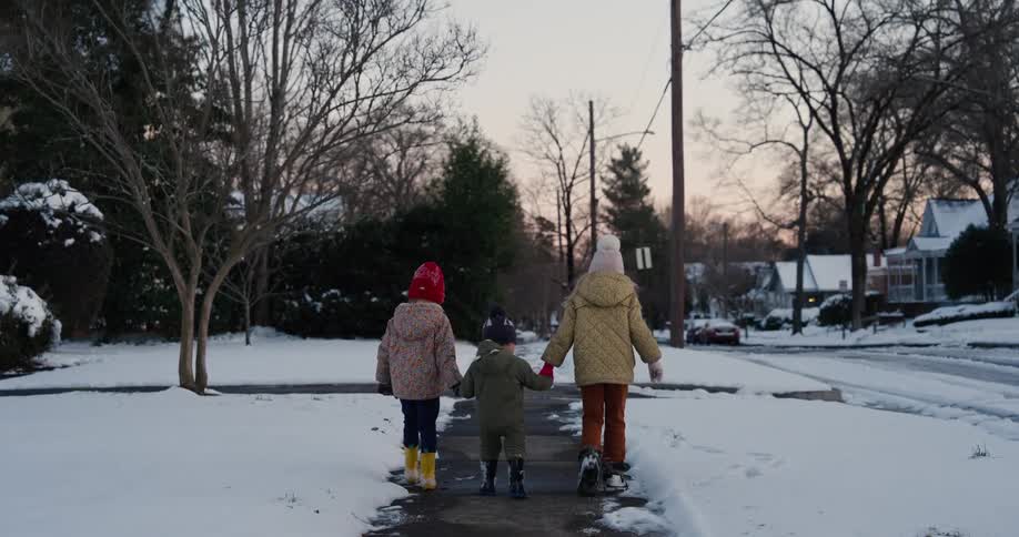 A view from behind shows three young children wearing winter coats and hats as they walk together along a snowy sidewalk in a quiet neighborhood during dusk