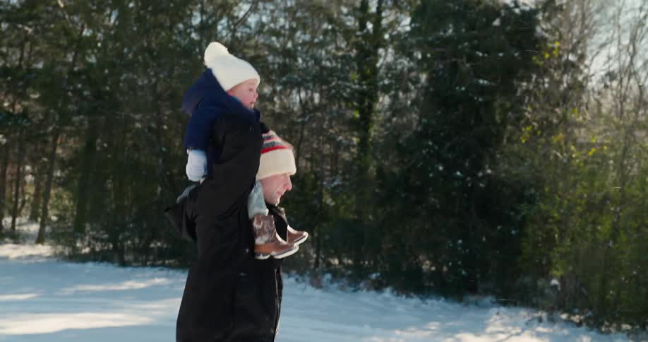 Father carries his little boy on his shoulders as they walk through the snow near the woods