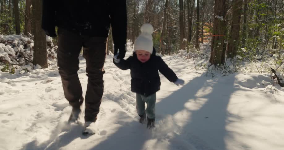 Child wearing a white beanie walking in the snow holding hands with an adult on a forest trail