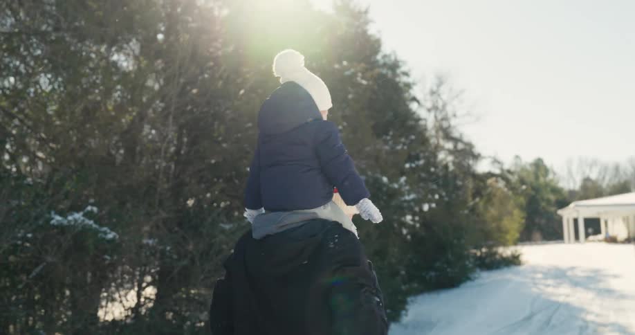 A young child wearing a blue coat and a white knit hat sits on the shoulders of an adult while moving through a snowy landscape with evergreen trees and a white building in the background during a bright winter day
