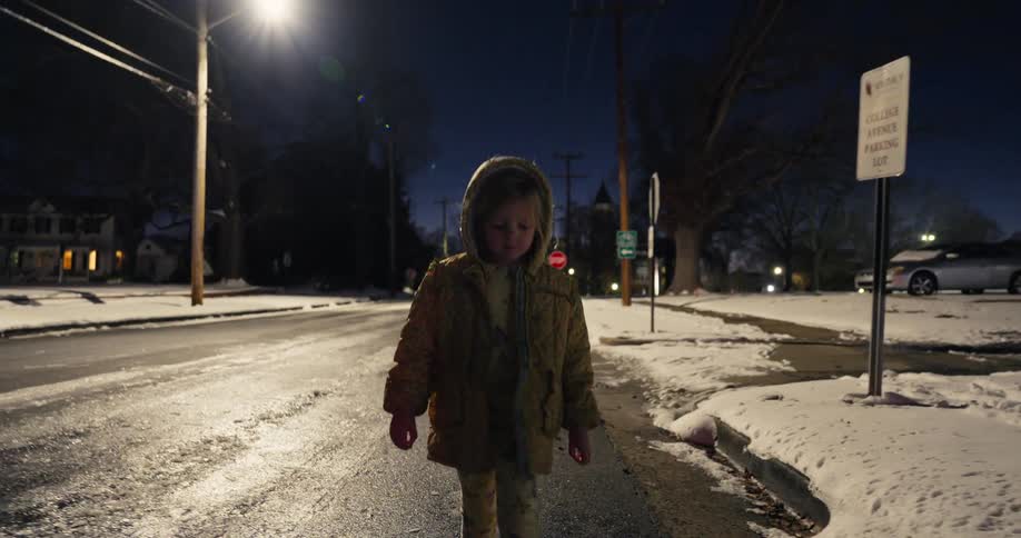 Little girl wearing a coat walking on a snowy street under a streetlight at night