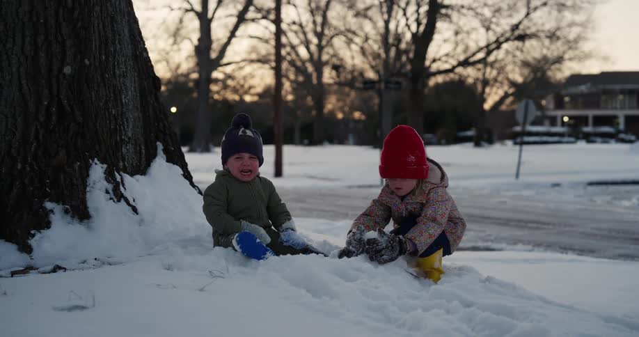 Two young children wearing winter jackets play with a large snowman in a snowy backyard during a winter day