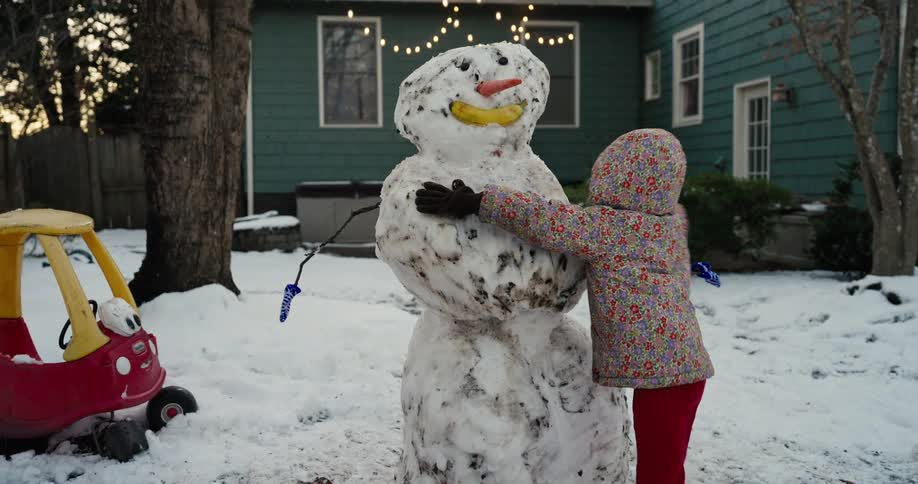 A young child wearing a floral jacket and red pants hugs a large snowman in a yard covered in snow during a winter day