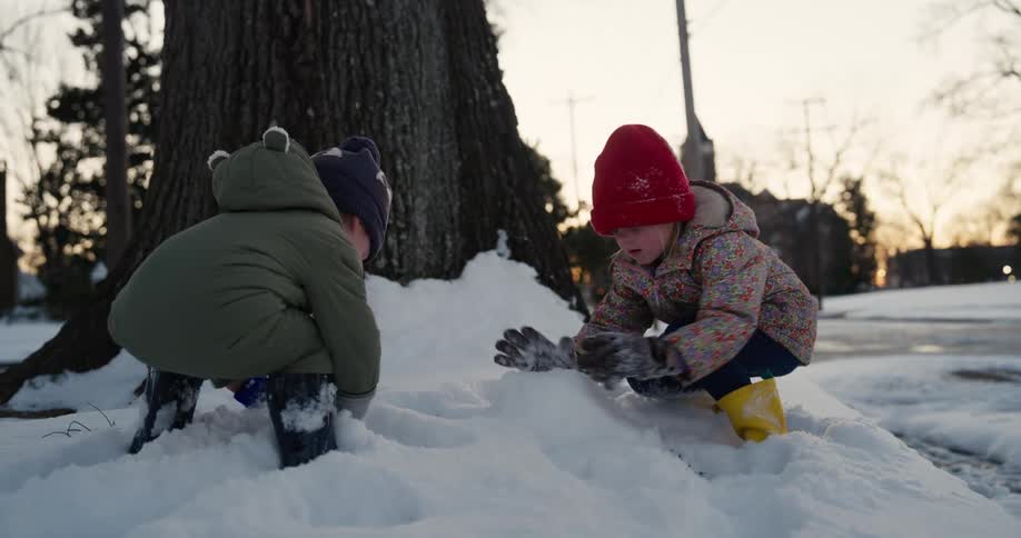 Two young children wearing winter jackets play with a large snowman in a snowy backyard during a winter day