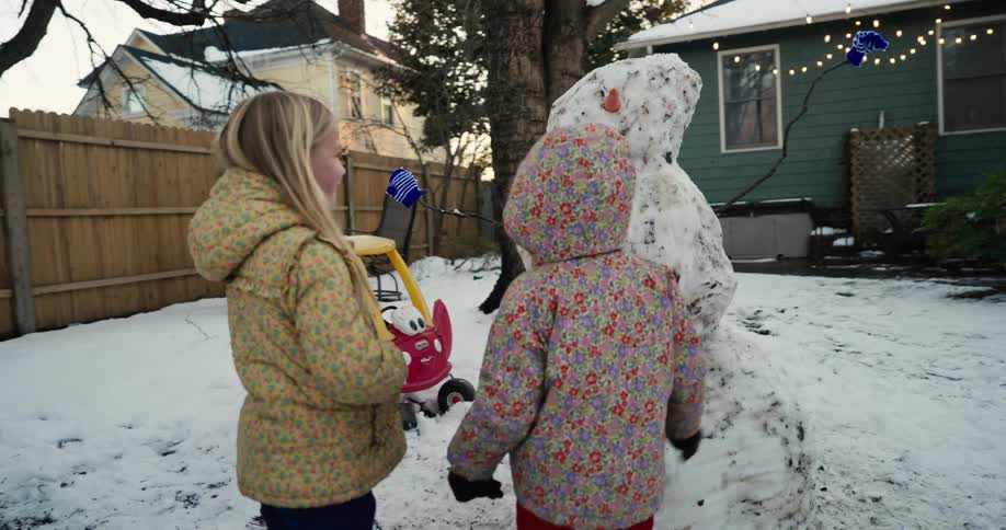Two young children wearing winter coats play with a tall snowman made of dirty snow in a backyard during a winter day