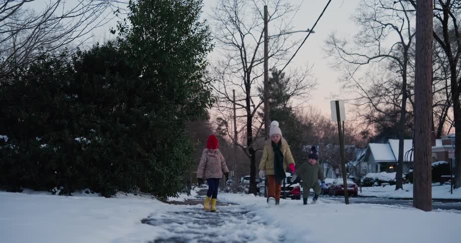 Three small children dressed in warm winter clothing walk toward the camera along a snow covered sidewalk in a quiet residential neighborhood during the late afternoon