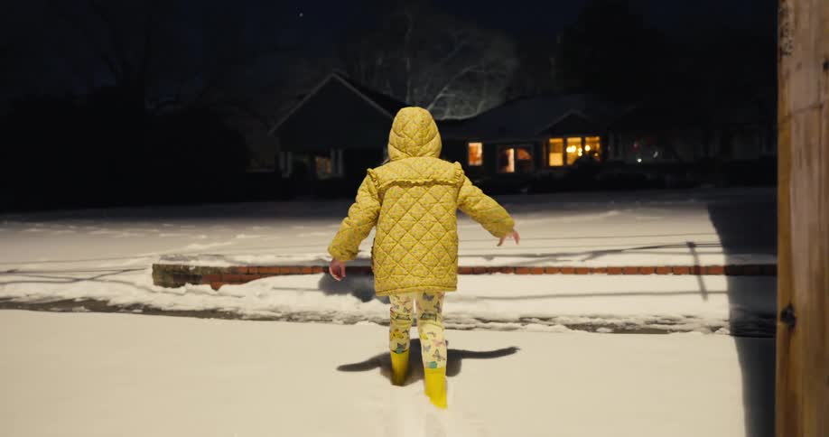 A young girl slowly walks through a snow covered neighborhood at night