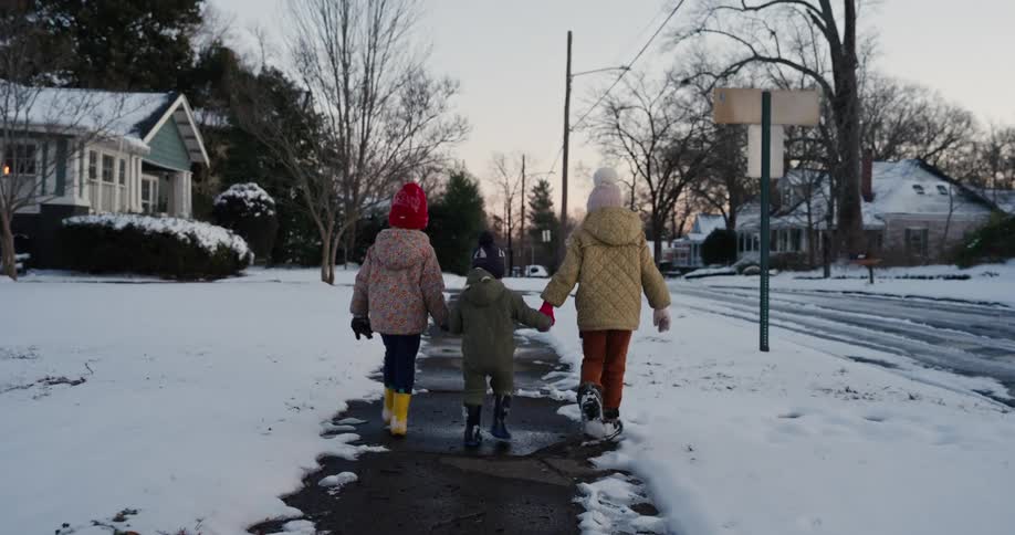 Three young children wearing colorful winter gear and boots hold hands while walking down a snow covered suburban sidewalk toward a street