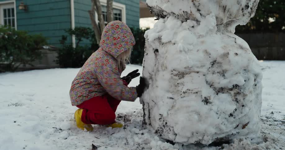 A child wearing winter clothing building a snowman on the front lawn of a building at midday