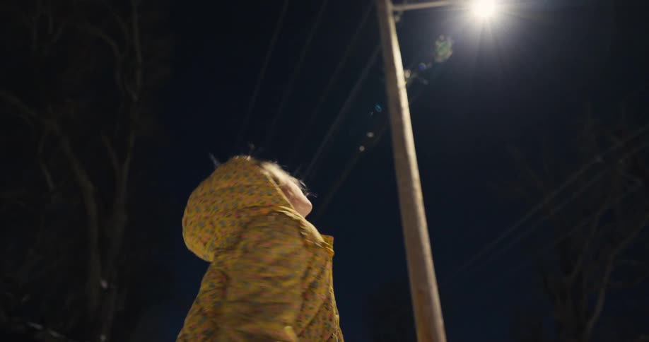 A young child wearing a yellow floral jacket with a hood looks up toward a bright street light in the dark night sky