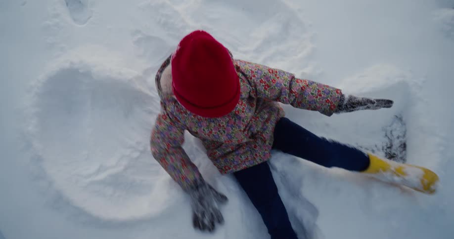 Little girl in a red hat lying in the snow making snow angels gets up and plays with another child