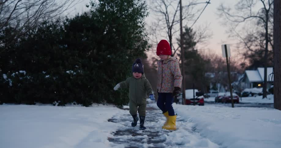 Two young children dressed in warm winter clothing and boots walk toward the camera on a narrow cleared path along a snow covered suburban sidewalk during a cold evening