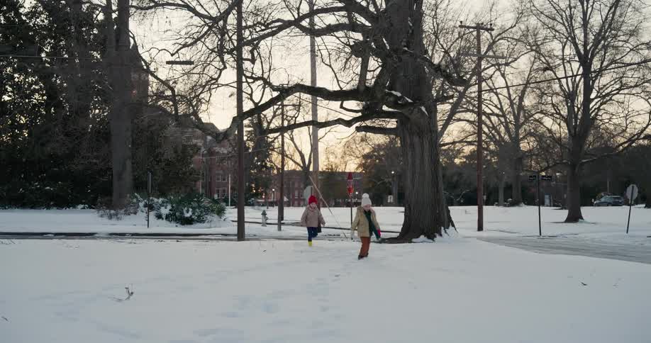 Two kids wearing winter clothing walking and playing in the snow on the sidewalk at midday