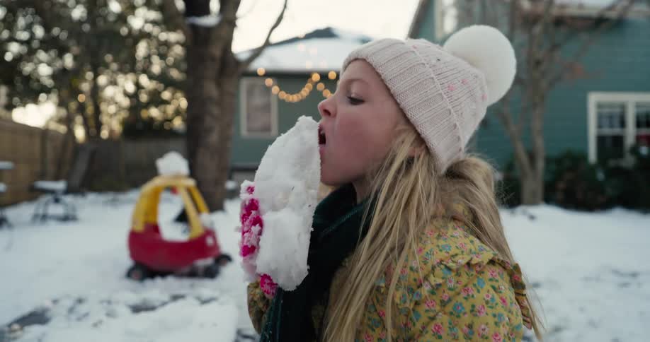 A young girl holds a chunk of snow and licks it while standing outdoors