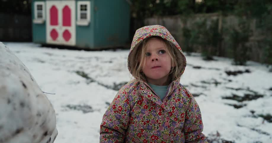 A small girl wearing a floral patterned coat with a hood stands in a snowy backyard and looks around while another girl walks past in the background near a blue shed
