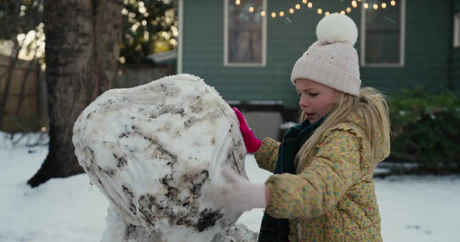 Girl wearing a white beanie shaping a snowman in a backyard with a house in the background