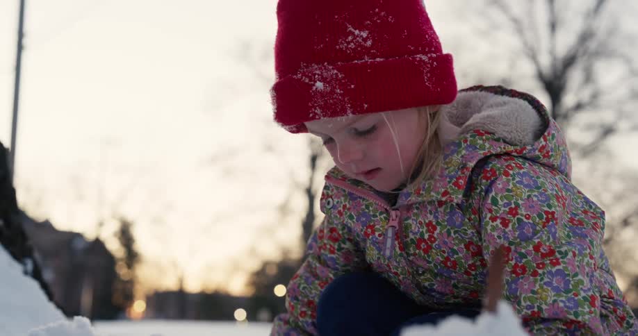 Little girl wearing a red beanie and floral coat playing in the snow at dusk