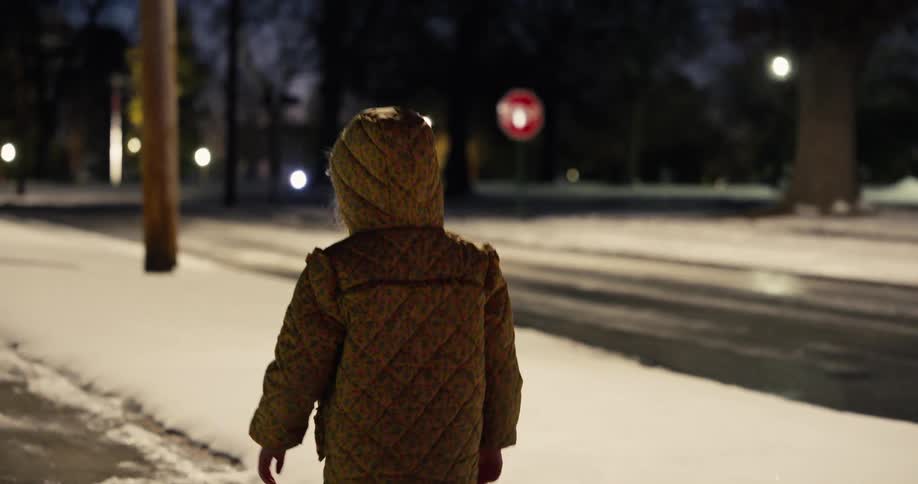 Little girl wearing a coat walking on a snowcovered sidewalk at night with a stop sign in the background