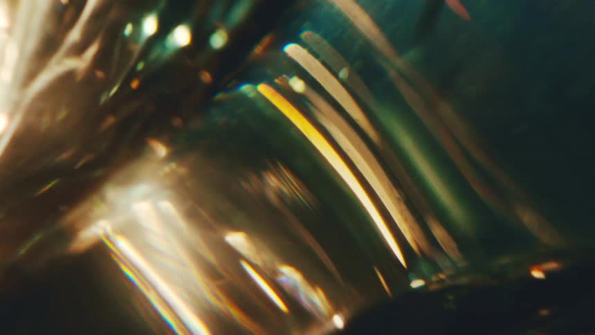 An extreme close view shows golden light reflecting and refracting through a glass material to create abstract glowing patterns and shimmering particles against a dark background
