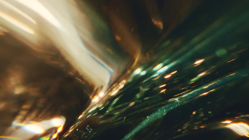 An extreme close view shows golden light reflecting and refracting through a glass material to create abstract glowing patterns and shimmering particles against a dark background