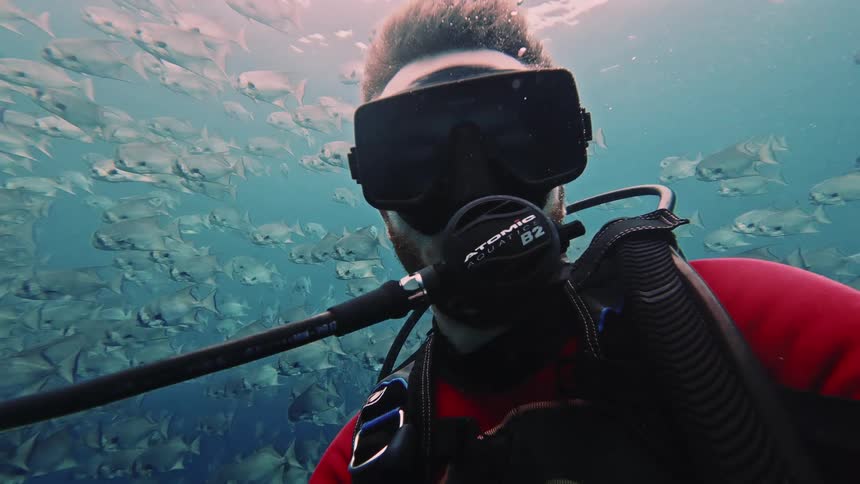 Underwater adult man scuba diving and filming himself next to a shoal of fish outdoors during day time