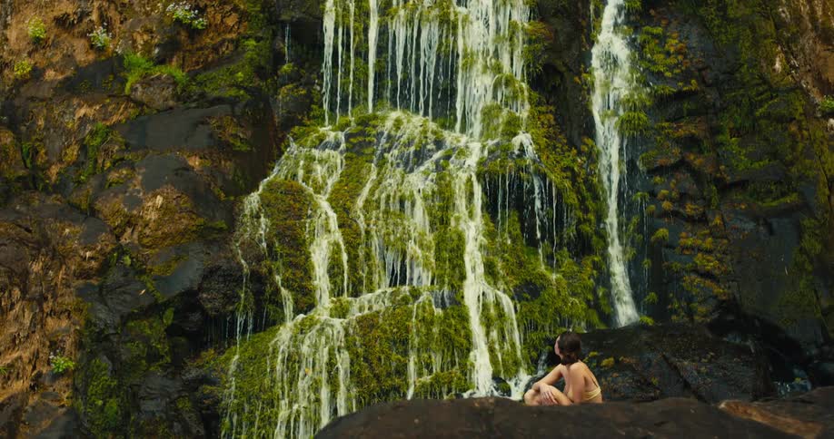Woman sits watching a waterfall flow over rocks on a mountain cliff